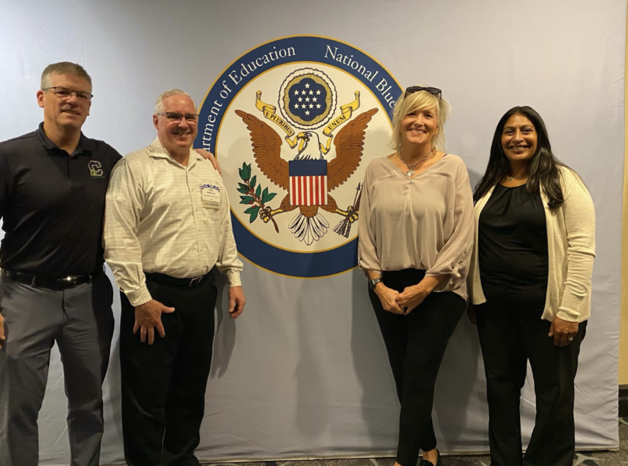 From left to right, Dr. Rob Bridges, Mr. Mark Matthews, Mrs. Julie Barthel, and Dr. Aarti Brooks smile in front of the National Blue Ribbon logo.